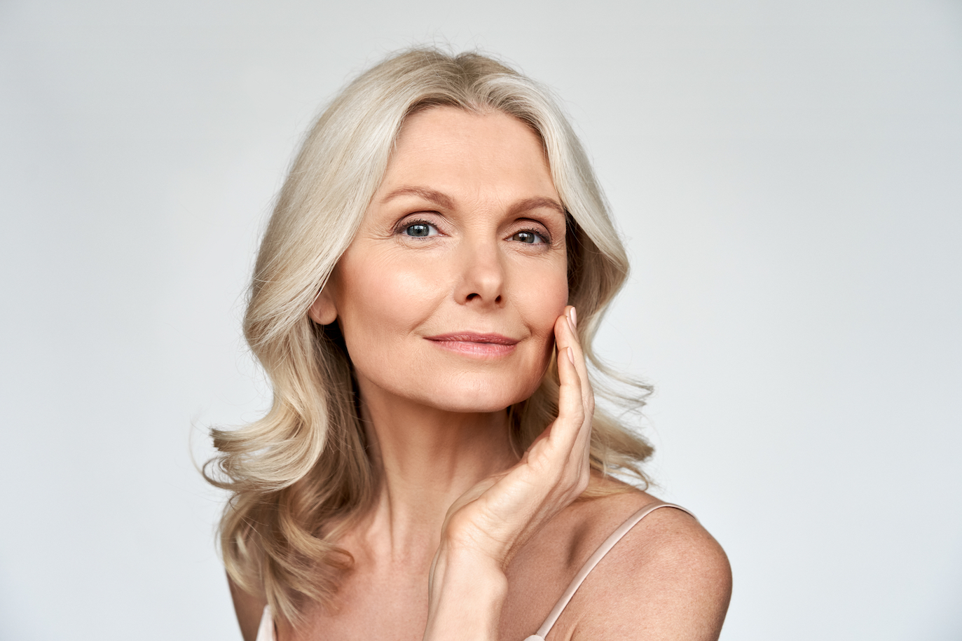 Woman in mid-50's with healthy skin looking at camera on white background. Her left hand is by her face to accentuate her skin's appearance